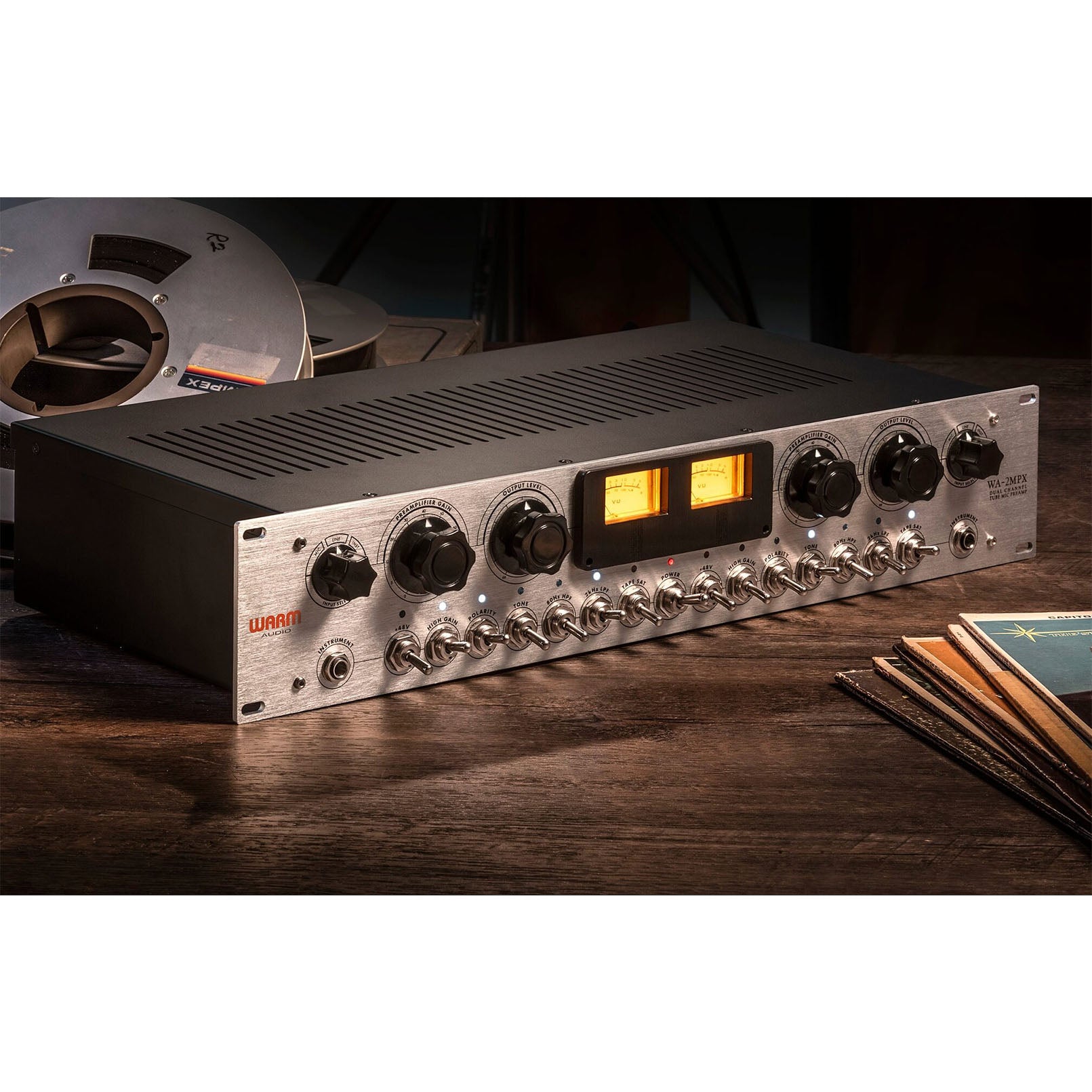 Warm Audio WA-2MPX 2-channel Tube Mic/Line/Instrument Preamp, angled view with vintage meters showing instrument input, input selector, gain and output level knobs. Also featuring switches for power, phantom power, high gain, polarity, tone, high and low pass filters and tape saturation. Placed on a wooden floor with Ampex tape rolls in the background.